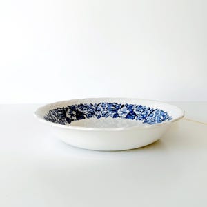 Può includere: Ciotola da portata in ceramica bianca con un motivo floreale blu sul bordo. La ciotola ha un bordo smerlato ed è posta su uno sfondo bianco. Il design floreale presenta fiori e foglie blu dettagliati.