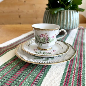 Può includere: Un set da tè in porcellana bianca con un design floreale. La tazza ha un bordo dorato e un delicato motivo floreale con fiori rosa e foglie verdi. Il piattino ha un design abbinato e un bordo ondulato.