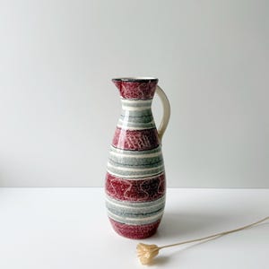 May include: A ceramic pitcher featuring a tapered neck and a curved handle. The exterior is decorated with horizontal bands of speckled deep red and muted sage green, accented by thin white lines and subtle, repeating wavy patterns throughout.