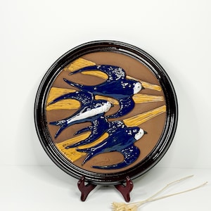 May include: A circular ceramic wall plaque depicting three blue swallows in flight against a brown backdrop with yellow accents. The birds have white markings and are framed in dark brown.