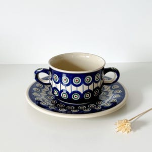 Può includere: Una tazza da tè in ceramica blu e bianca con due manici e un piattino abbinato. La tazza e il piattino presentano un motivo ripetuto di cerchi blu, verdi e rossi. La tazza da tè è appoggiata sul piattino.