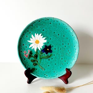 May include: A decorative turquoise ceramic plate with a floral design. The plate features a white daisy, a dark blue flower, and small pink flowers. The plate is displayed on a dark red stand.