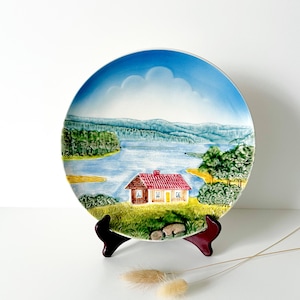 May include: A decorative ceramic plate featuring a scenic landscape with a house, lake, and trees. The plate has a blue sky with clouds and is displayed on a dark red stand. The house has a red roof and yellow door.