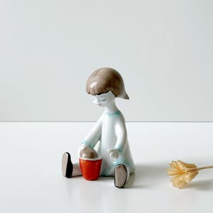 Può includere: Una statuetta in porcellana di un bambino seduto con capelli castani, che indossa un vestito bianco con accenti azzurri. Il bambino tiene in mano un piccolo secchiello arancione brillante con l'interno marrone. La figura ha scarpe nere e finitura liscia.