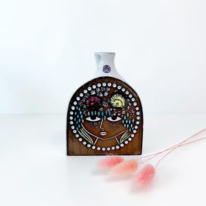 May include: A brown ceramic vase with a white neck and a painted face design. The face has large eyes, a small nose, and a detailed floral crown. The vase is decorated with white dots and diamond shapes.