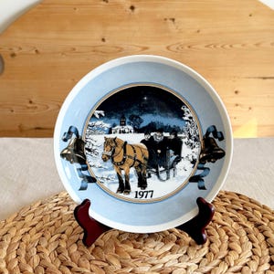 May include: A decorative plate with a winter scene featuring a horse-drawn sleigh, bells, and the year 1977. The plate has a light blue border and a dark blue center with a snowy landscape. The plate is displayed on a wooden stand.