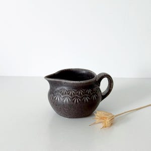 May include: A small, dark gray ceramic creamer with a speckled finish and a rounded body. The exterior features a repeating embossed fan-shaped pattern around the center. It has a curved handle on one side and a small pouring spout on the other.