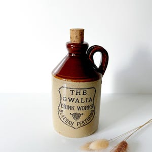 Op de afbeelding: Een bruine en beige stenen kruik met een handvat en een kurken stopper. De kruik heeft de tekst "THE GWALIA DRINK WORKS BLAENAU FESTINIOG" in het zwart. Deze vintage kruik is waarschijnlijk voor het bewaren van drank.