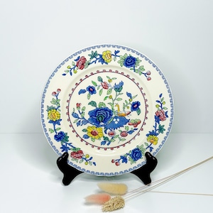 Op de afbeelding: Een crèmekleurige decoratieve plaat met een bloemenontwerp. De plaat heeft een centraal motief van blauwe bloemen, gele accenten en rode en roze bloemendetails. De rand heeft een blauw-wit patroon. De plaat wordt op een zwarte standaard gepresenteerd.