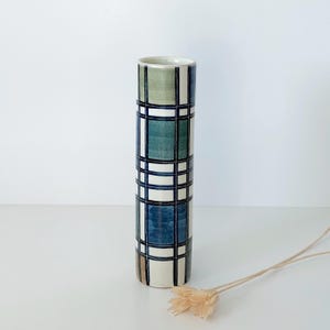 May include: A tall, cylindrical ceramic vase with a plaid pattern in shades of green, blue, and white. The vase has a smooth, glossy finish and a narrow opening. The design is a grid of dark blue lines over the colored sections. A decorative item for the home.