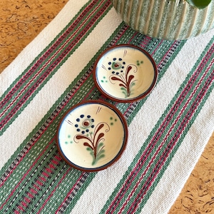 Puede incluir: Dos platos pequeños de cerámica redondos con fondo blanco y diseños florales marrones, verdes y azules. Los platos están sobre un mantel a rayas blancas y verdes.