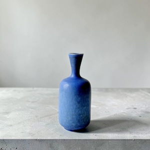 Op de afbeelding: Een kleine, cilindervormige keramische vaas met een smalle hals en een uitlopende rand. De vaas heeft een gevlekt, matblauw glazuur met subtiele kleurvariaties, wat zorgt voor een getextureerd uiterlijk over het gehele oppervlak.