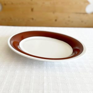以下が含まれることがあります： 白いセラミック製のプレートで、幅広の茶色の縁取りが施されています。プレートは白いテーブルクロスの上に置かれています。