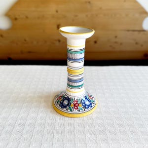 Könnte beinhalten: Ein weißer Kerzenhalter aus Keramik mit einem gelben, blauen und grünen Streifenmuster und floralen Akzenten. Die Basis des Halters ist mit bunten Blumen verziert.