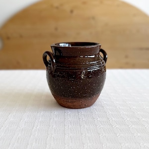 Op de afbeelding: Een kleine, bruine aardewerken pot met twee handvatten. De pot heeft een gespikkelde glazuur en een licht afgeronde vorm.
