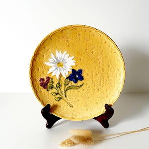 May include: A yellow ceramic plate with a floral design featuring a white daisy, a blue flower, and red berries. The plate has a scalloped edge and is displayed on a dark brown stand. The plate's diameter is approximately 10 inches.