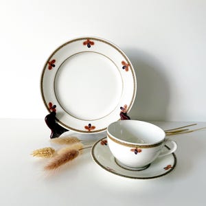 May include: A white porcelain teacup and saucer set with a matching dinner plate. The set features a gold border with a repeating pattern of red and blue floral accents. The plate is propped up on a dark wooden stand.