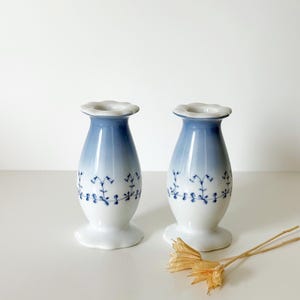 May include: A pair of white porcelain candle holders with a blue gradient finish at the top and a delicate blue floral vine pattern encircling the midsection. Each holder features a scalloped rim and a flared base for stability.