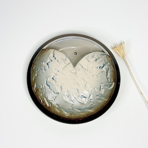 May include: A handmade ceramic heart-shaped dish with a dark brown rim and a light gray interior. The heart-shaped section has a textured, off-white surface with blue accents. A dried flower stem is to the side.