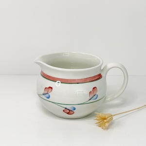 May include: White ceramic creamer with a red and green band around the middle. The creamer features a floral design with red and blue flowers and green stems. It has a handle and a spout.