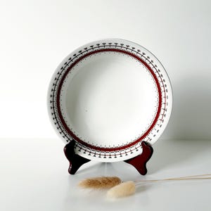 May include: White ceramic plate with a decorative red and black border, displayed on a dark wooden stand. The plate features a simple, yet elegant design, suitable for serving food. Dried decorative plants are in the foreground.