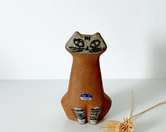 May include: A ceramic cat figurine in shades of brown, with black and white details. The cat has a stylized face and striped paws. A blue and white sticker is affixed to the base. The figurine is a Lisa Larson design.
