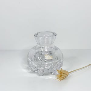 Peut inclure: Un vase à bourgeon en verre transparent avec un corps arrondi texturé et un dessus évasé. Le vase a une forme unique et organique et repose sur une surface blanche. Une tige de fleur séchée se trouve à proximité.