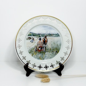 May include: A decorative white porcelain plate with a gold rim and a pastoral scene. The plate features a detailed illustration of a horse-drawn carriage harvesting a field. The border has a repeating floral design.