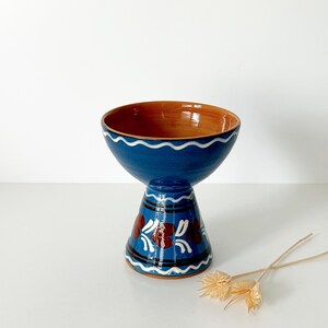 May include: A blue ceramic bowl with a brown interior, supported by a pedestal base. The bowl has a wavy white trim. The pedestal is decorated with red floral designs and black stripes. A decorative piece.