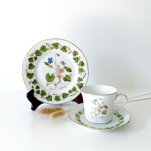 May include: A white porcelain teacup, saucer, and plate set. The set features a botanical design with green leaves, purple and blue flowers, and gold trim. The plate is 17.78 cm in diameter.