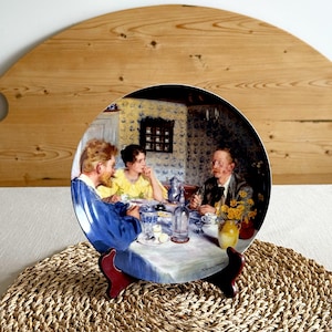 May include: Decorative plate featuring a painting of three people seated at a table, with a white tablecloth and various dishes. The plate is displayed on a dark red stand, set against a woven mat and a wooden background.
