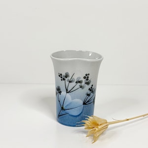Può includere: Un vaso in ceramica con una sfumatura azzurra e bianca. Il vaso presenta un motivo floreale nei toni del blu e del nero. Il vaso ha un bordo ondulato ed è alto circa 13 cm.