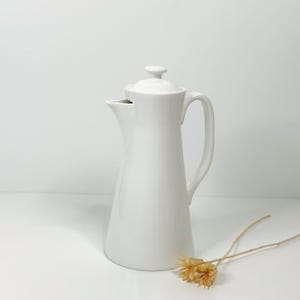 Op de afbeelding: Een witte keramische koffiepot met een deksel en een handvat. De pot heeft een slanke, conische vorm en een tuit om uit te schenken. Het handvat is gebogen en het deksel heeft een kleine knop. Een gedroogde bloem staat op de voorgrond.
