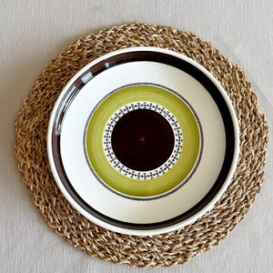 May include: A decorative ceramic plate with a brown, white, and green concentric circle design. The plate sits on a round, woven placemat. The plate's center is dark brown, surrounded by a green band, a white band, and a brown rim. The placemat is made of natural fibers.