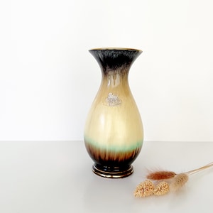 May include: A tall, decorative vase with a flared top and a rounded body. The vase features a gradient of colors, transitioning from black at the top to tan, green, and brown at the base. A gold band accents the rim and base. A small emblem is visible on the vase.