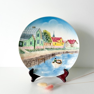 May include: A decorative ceramic plate featuring a village scene with colorful houses, a river, and a boat. The plate has a blue sky background and is displayed on a dark brown stand. The plate's design includes green, yellow, and red houses.