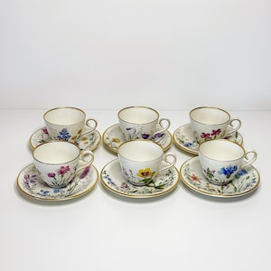 Puede incluir: Seis tazas y platillos a juego. Cada taza y platillo presenta un diseño floral en varios colores, incluyendo rosa, amarillo y azul, con ribetes dorados. Las tazas son blancas con borde dorado.