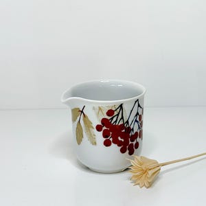 Può includere: Una lattiera in ceramica bianca con beccuccio, decorata con un motivo di bacche rosse e foglie marroni. La lattiera ha un corpo arrotondato e un piccolo manico. Un fiore secco è in primo piano.