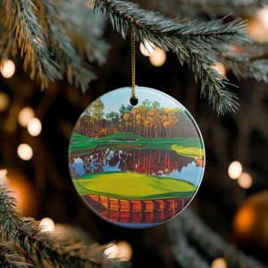 May include: Round ceramic Christmas ornament featuring a golf course scene with a lake and trees. The ornament is hanging from a gold string against a blurred background of Christmas lights and a pine branch.