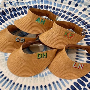 May include: Four straw sun visors with different embroidered initials: AH, CB, KF, and LN. The visors are brown and have a wide brim.