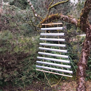 Può includere: Un carillon in argento con otto tubi appesi a un bastone di legno e una corda. Il carillon è appeso a un ramo di albero.