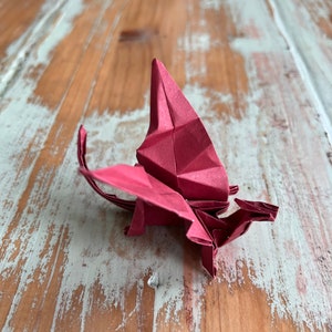 May include: A red origami dragon with wings spread out, sitting on a light brown wooden surface with white paint.