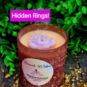 May include: A brown glass candle jar with a frosted finish. The candle is a light orange color with a purple flower on top. The jar has a white label with the text "Homemade SONshine Bamboo Coconut Mystery Crystal Ring Size & Natural Soy 1 oz" on it. The candle is sitting on a bed of gold glitter.
