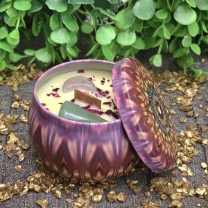 May include: A decorative candle in a purple and pink patterned tin, with a mandala-patterned lid. The candle has a light yellow wax, a wooden wick, and is decorated with rose petals and crystals. The candle is surrounded by gold-colored decorative stones.