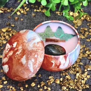 May include: A decorative candle in a round tin with a lid. The candle is pink with two green butterfly accents and a wooden stick. The lid and tin have a red, white, and blue marbled design. A black stone sits inside the tin.