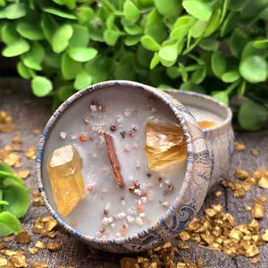May include: A decorative candle in a blue and white patterned tin. The candle is light blue with crystals, small stones, and a cinnamon stick. The candle is surrounded by gold-coloured stones and green foliage.
