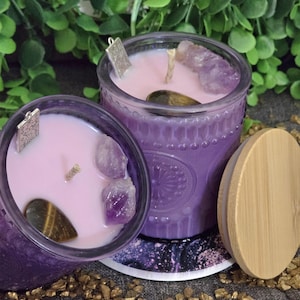 May include: Two purple glass candles with pink wax, adorned with amethyst crystals and a tiger's eye stone. Each candle has a silver charm and a wooden wick. A wooden lid sits beside one candle.