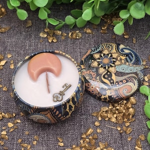 May include: A decorative candle in a round tin with a detailed, colourful paisley design. The candle has a crescent moon and key charm. The lid is off, revealing the white candle wax. Gold-coloured decorative stones and green leaves surround the candle.