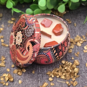 May include: A decorative candle in a patterned tin with a lid. The candle has a creamy wax and is decorated with red stones and a cinnamon stick. The tin is open, showing the candle's contents.