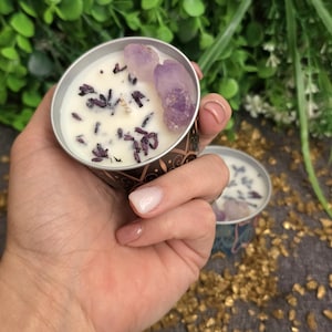 May include: A small, round candle in a decorative tin. The white wax is adorned with dried lavender and two amethyst crystals. The tin has a patterned design. The candle is held in a hand.
